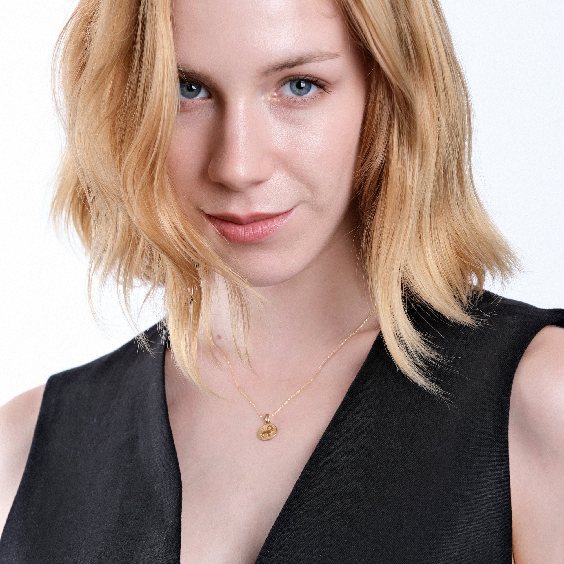 Blonde woman with blue eyes wearing black top and gold necklace, smiling confidently against white background.