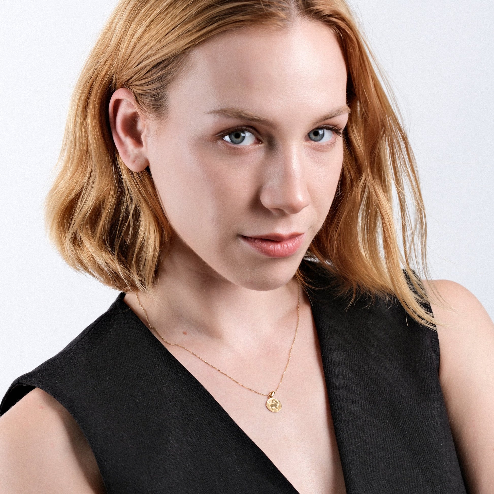 Blonde woman wearing black top and gold necklace, posing confidently with subtle smile on white background.
