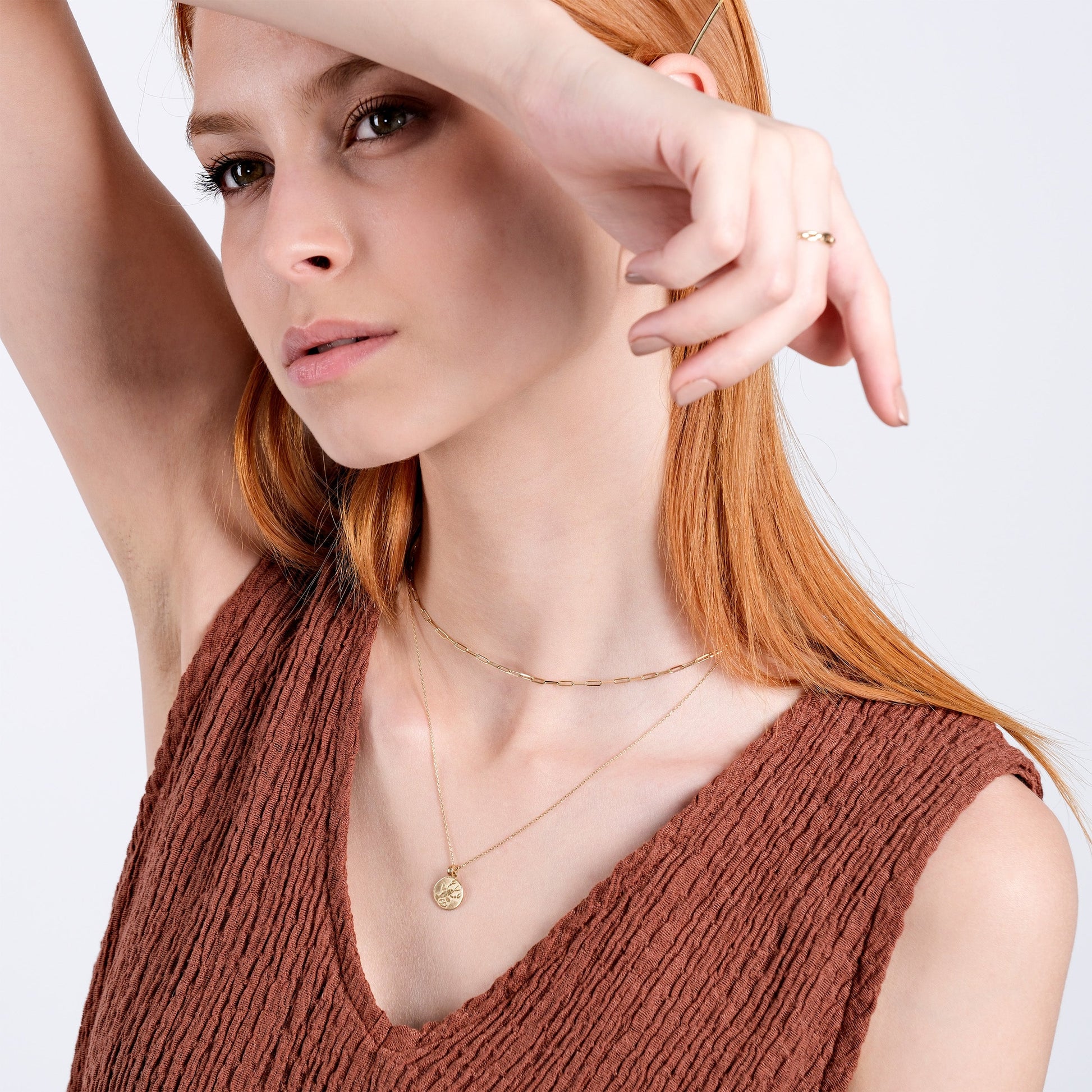 Woman with red hair wearing gold necklaces and brown textured top, posing with hand near face.