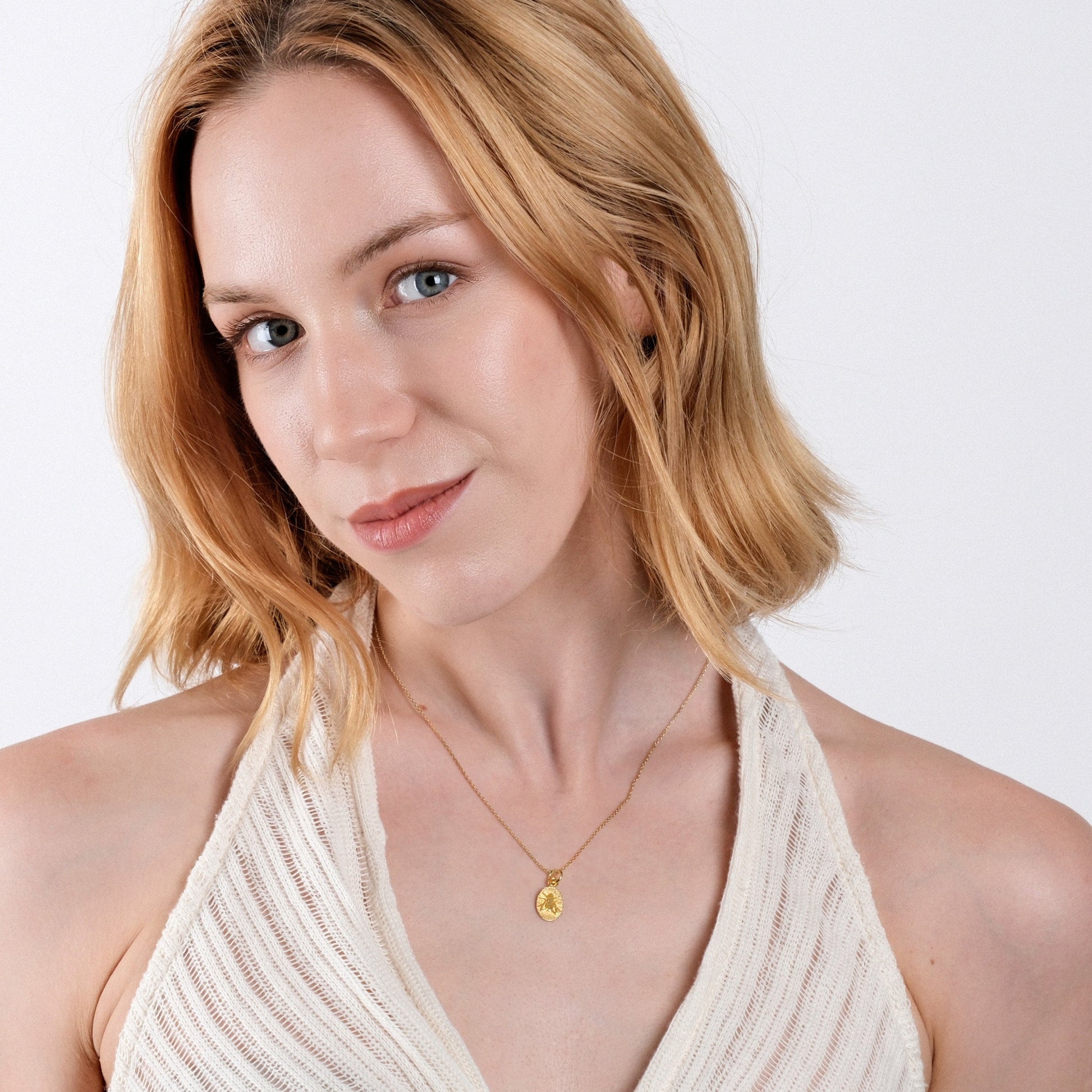 Woman with blonde hair wearing a gold necklace and white halter top, smiling softly against a plain background.