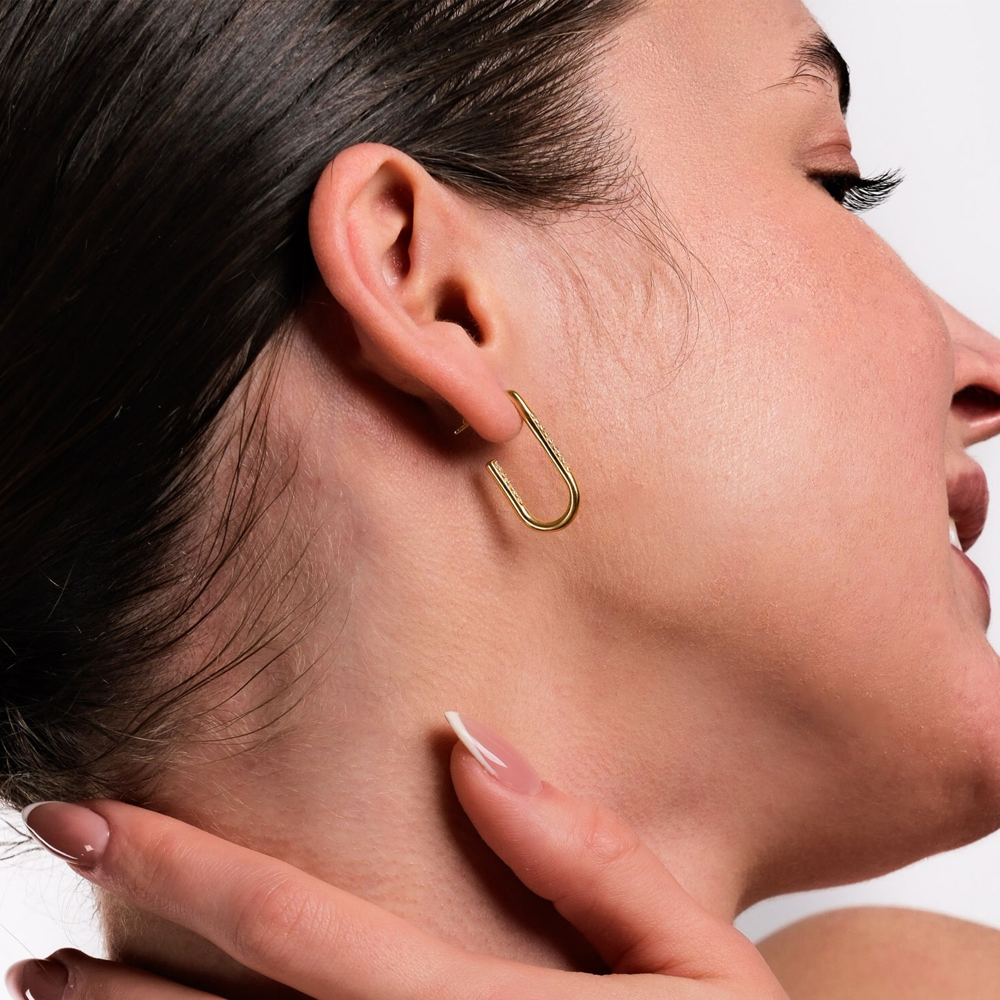 A close-up side view of a woman wearing 18K Gold Vermeil Geometric Earrings in a safety pin shape with sparkling stones. Her dark hair is pulled back, highlighting the modern and elegant design against a neutral background.