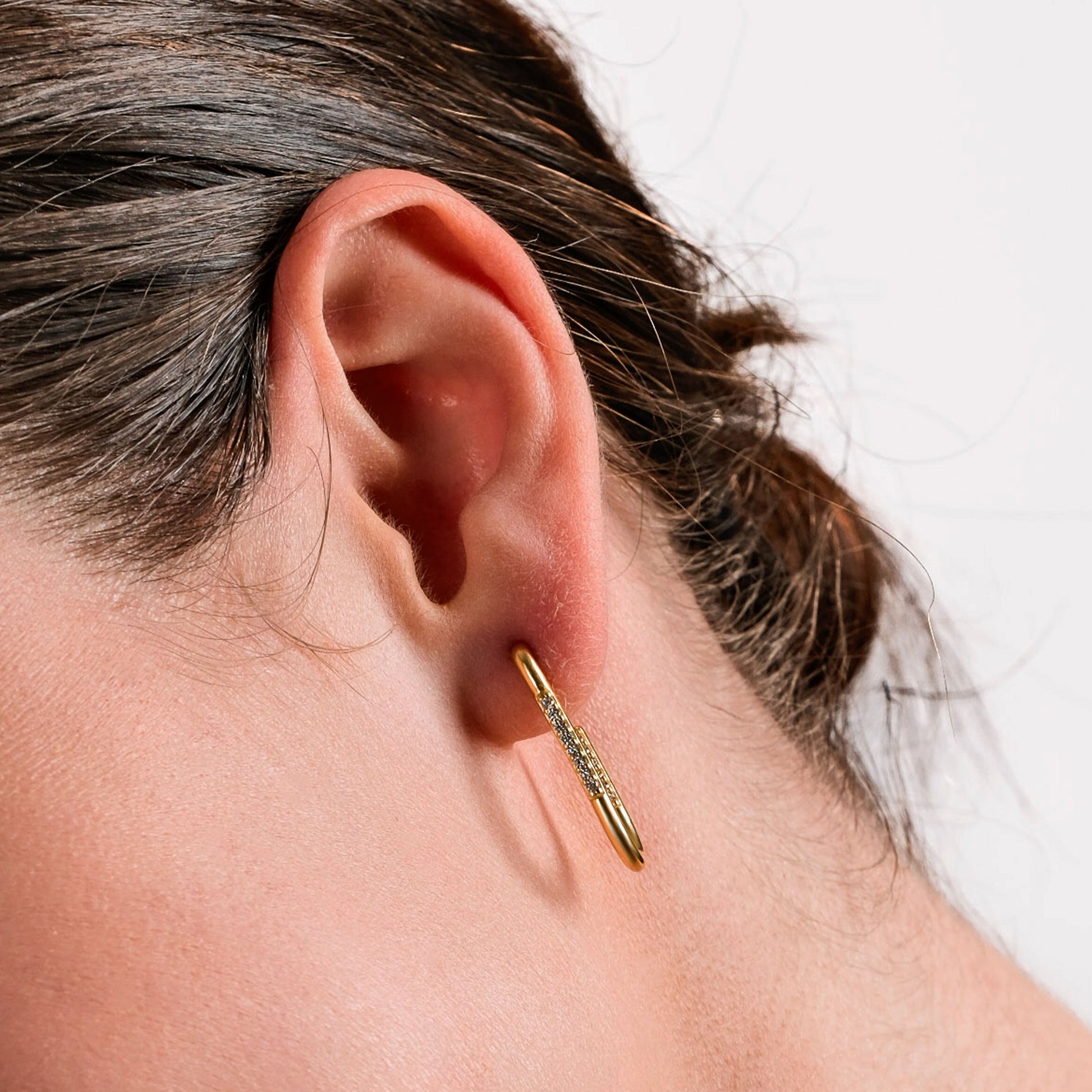 A close-up of a woman's ear adorned with a gold safety pin-shaped earring encrusted with small sparkling stones. The earring is positioned through the earlobe, creating a unique and modern jewelry style. The woman's dark brown hair is neatly pulled back, highlighting the earring against her fair skin. The background is a clean, neutral white, emphasizing the details of the jewelry.
