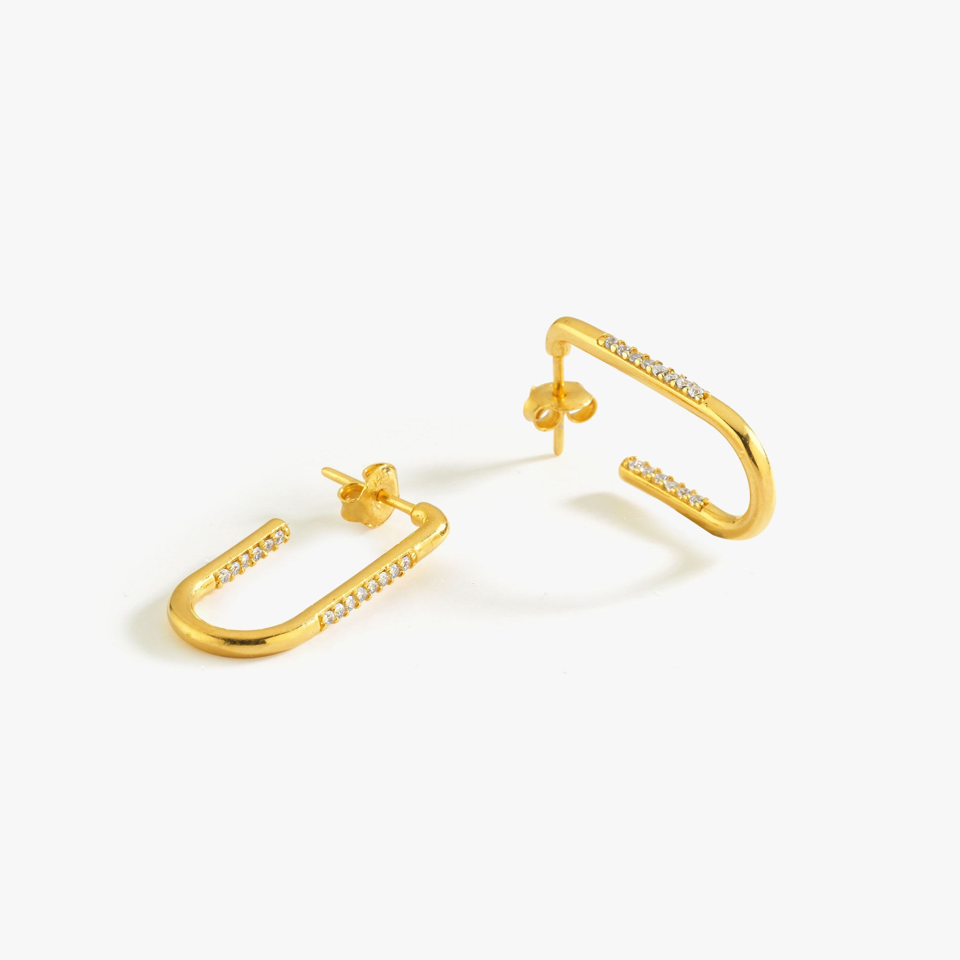 A pair of gold-plated safety pin-shaped earrings with sparkling cubic zirconia stones, displayed on a clean white background. The earrings have a modern and minimalist design, with a curved open-ended structure and a secure butterfly back closure. The small stones are set along the front-facing edges, adding a subtle touch of elegance and shine.