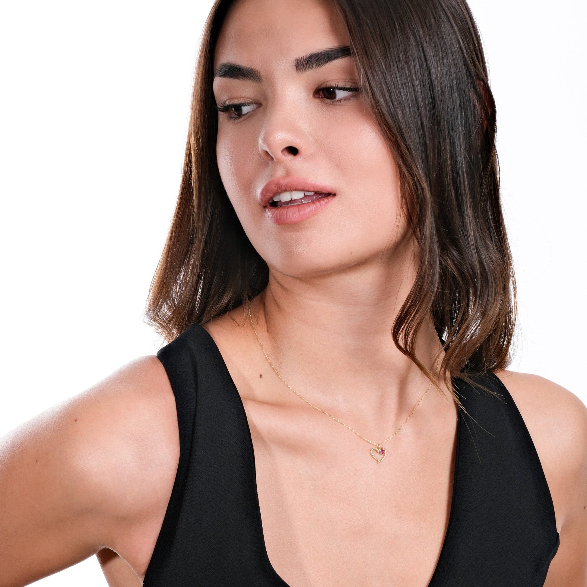 Woman wearing elegant black dress and heart-shaped necklace, looking sideways against white background.