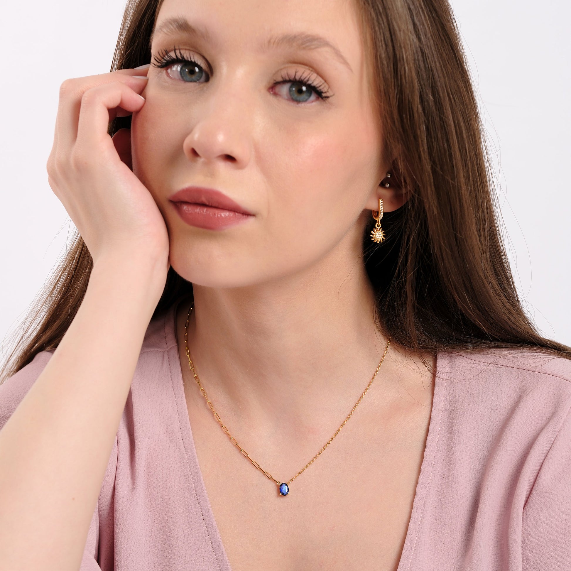 Woman wearing elegant gold jewelry, blue pendant necklace, sunburst earrings, soft expression, light pink blouse.