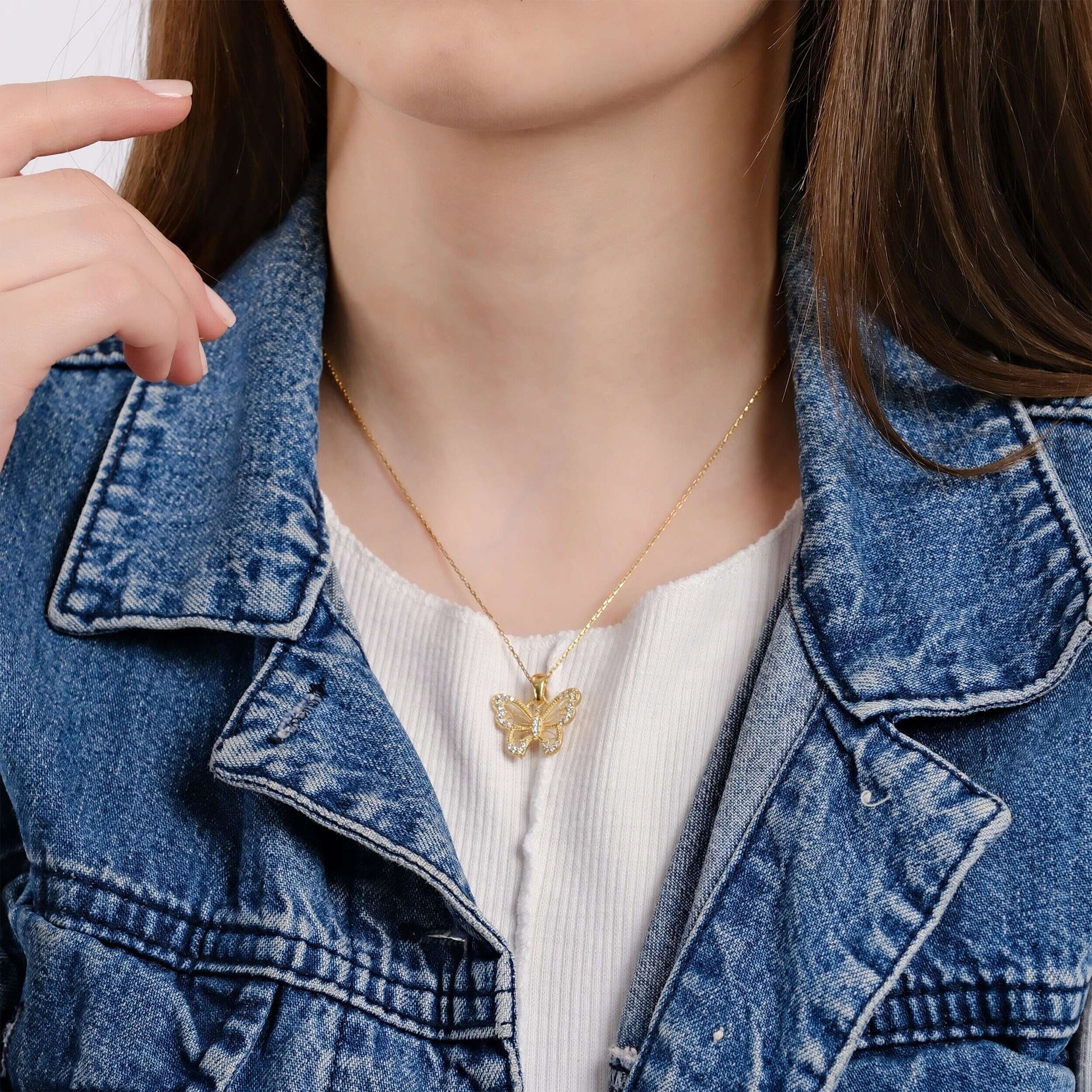 Close-up view of a woman's neck area, featuring an 18K gold vermeil butterfly necklace on a thin gold chain, set against a white top and a blue denim jacket.
