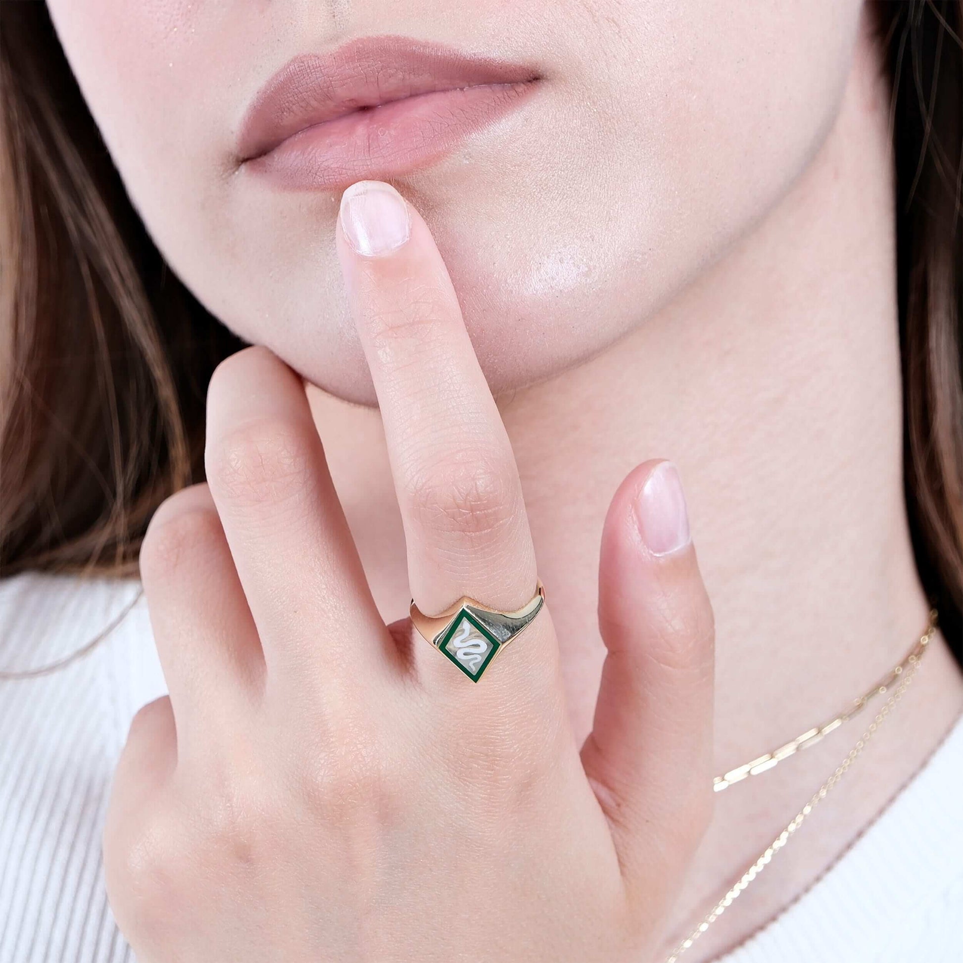 A close-up of a woman's face, focusing on her lips and chin, with her hand gently touching her chin. She is wearing a gold snake signet ring with a green and white enamel snake design.