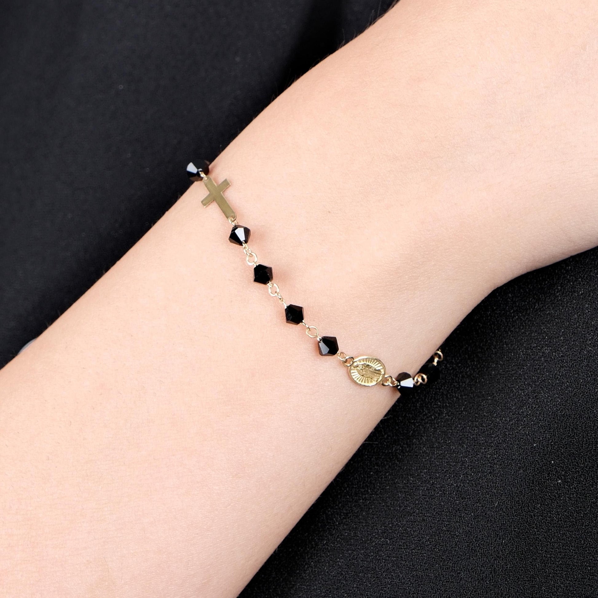 A close-up of a woman's wrist wearing a delicate 14K gold bracelet with black onyx beads, featuring a small cross charm and a medallion.