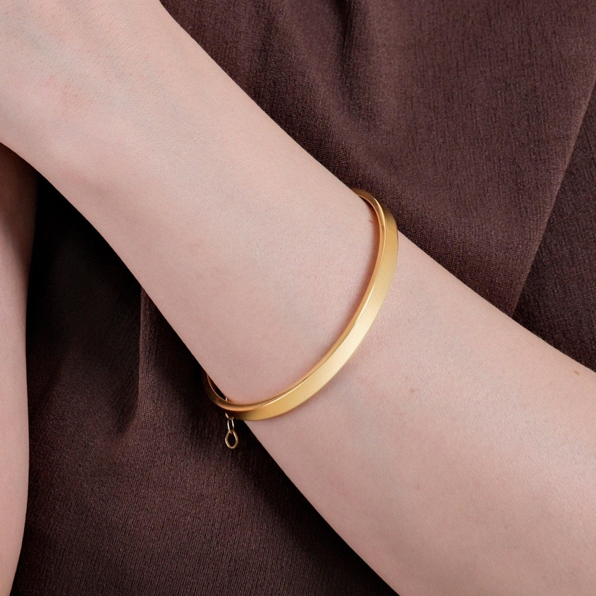 A close-up view of a person's arm wearing an 18K gold vermeil cuff bracelet against a brown fabric background.