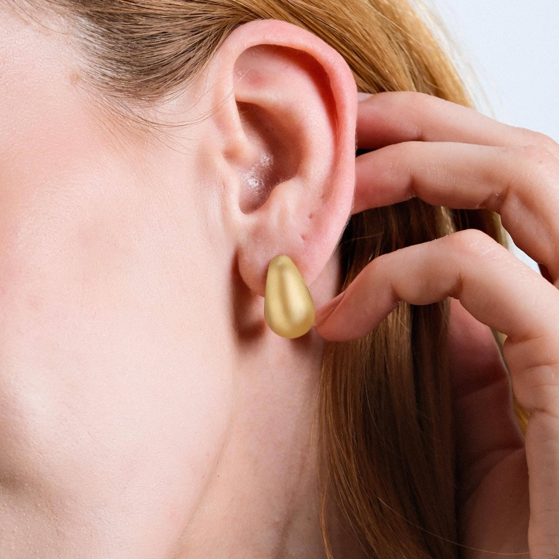 lose-up of a woman's ear with a single 18K gold vermeil tear drop earring. The matte gold finish of the earring contrasts with the smooth skin, as the woman touches her earring lightly.