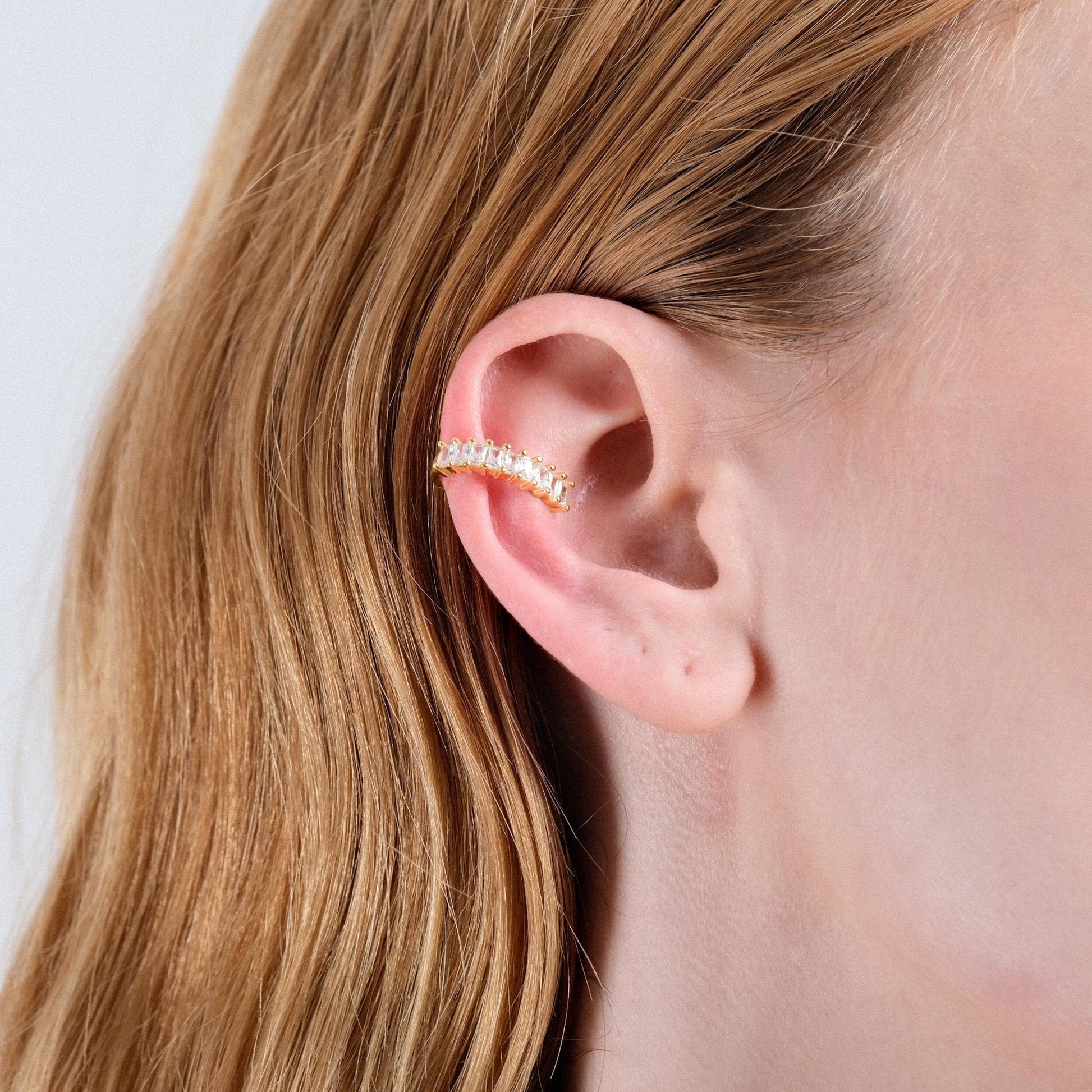A side view of a woman's ear adorned with a gold ear cuff featuring clear stones. Her light brown hair is tucked behind the ear, showcasing the cuff's placement.