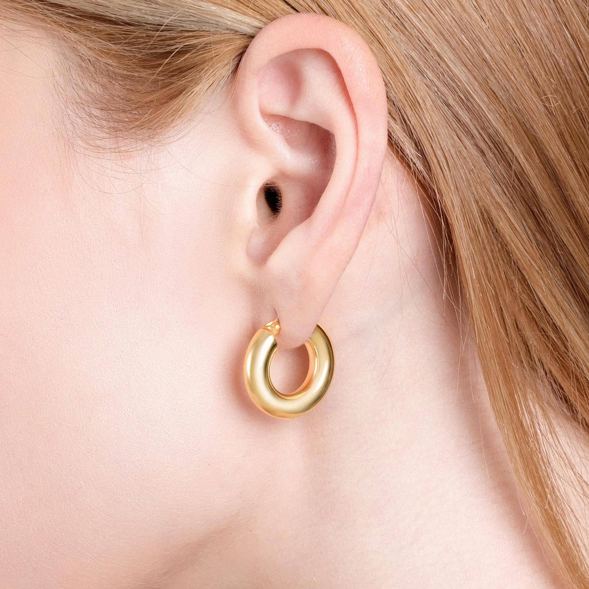 A detailed close-up of a person's ear showing a shiny, thick 18K gold vermeil chunky hoop earring, highlighting its smooth, rounded shape against light skin and blonde hair.