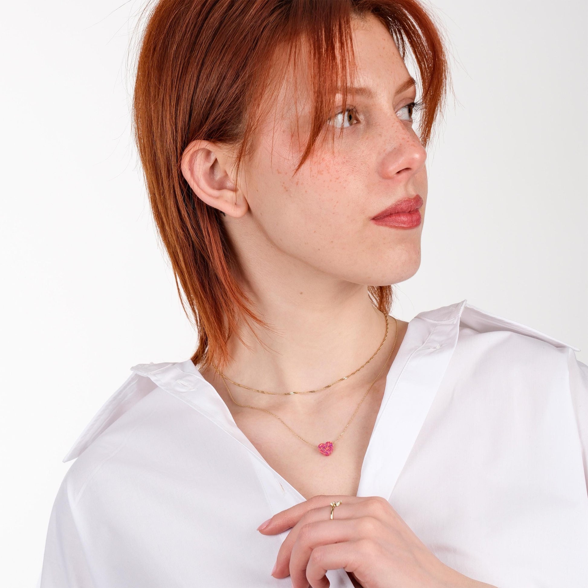 Woman looking to the side while wearing a layered 14K solid gold dainty opal heart necklace, paired with a classic white shirt and an elegant gold ring.
