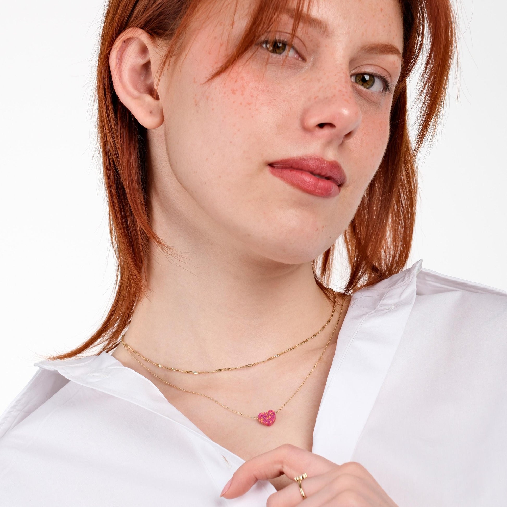 Red-haired woman glancing sideways while modeling a 14K solid gold dainty opal heart necklace and a dainty gold ring, styled over a crisp white shirt.