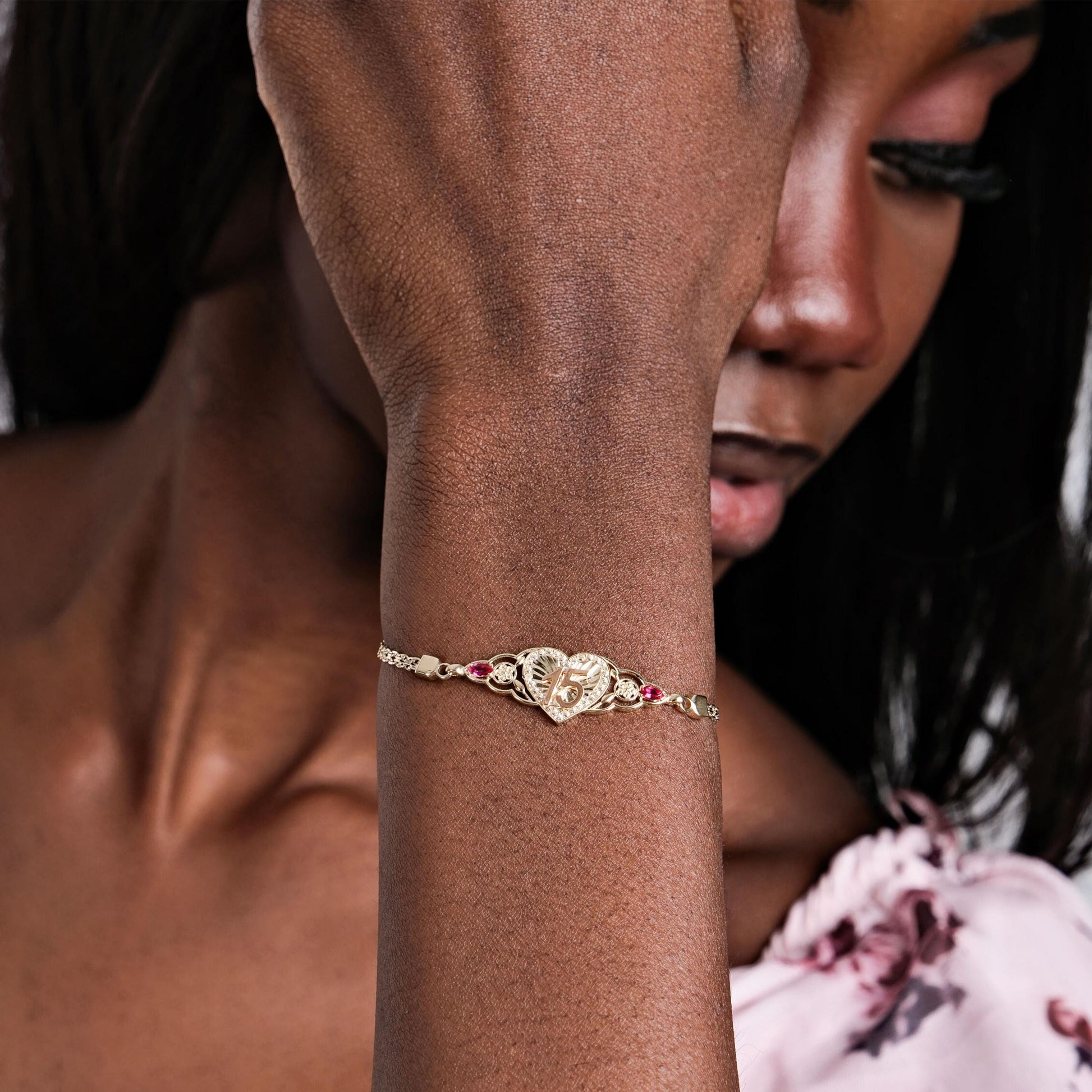 A woman's wrist adorned with a Quinceañera Gift bracelet featuring a gold heart centerpiece, delicate filigree, and red gemstones.