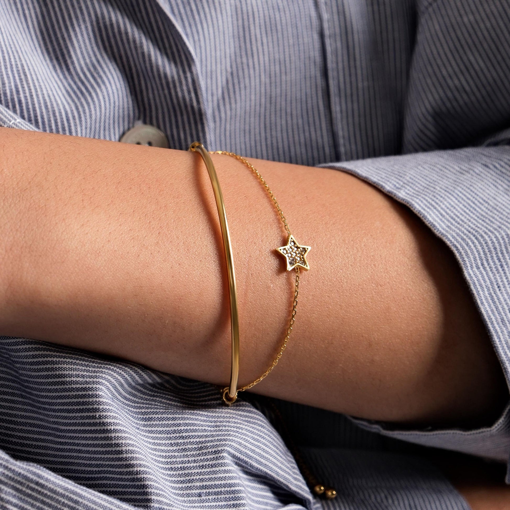 A woman wearing a blue pinstriped shirt showcases a delicate 18K gold vermeil sliding adjustable bracelet with a small star charm on her wrist.