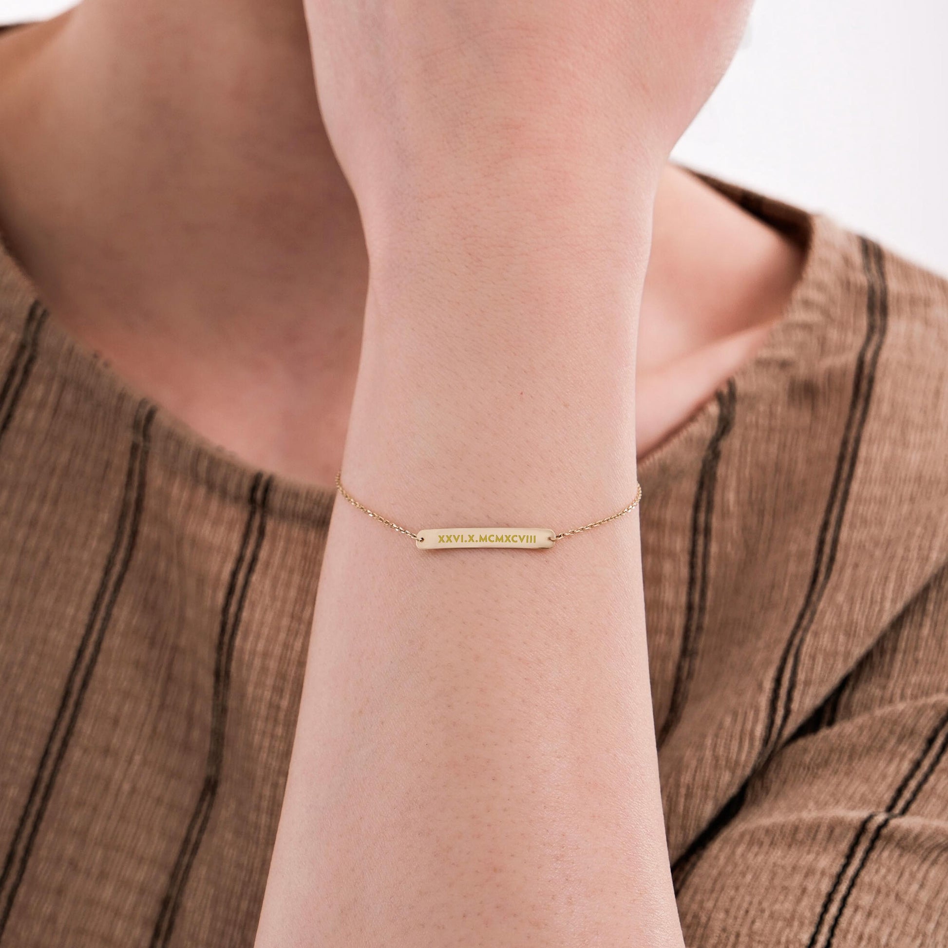Close-up of a woman's wrist wearing a 14K solid gold engraved bar bracelet with Roman numerals on a delicate chain.