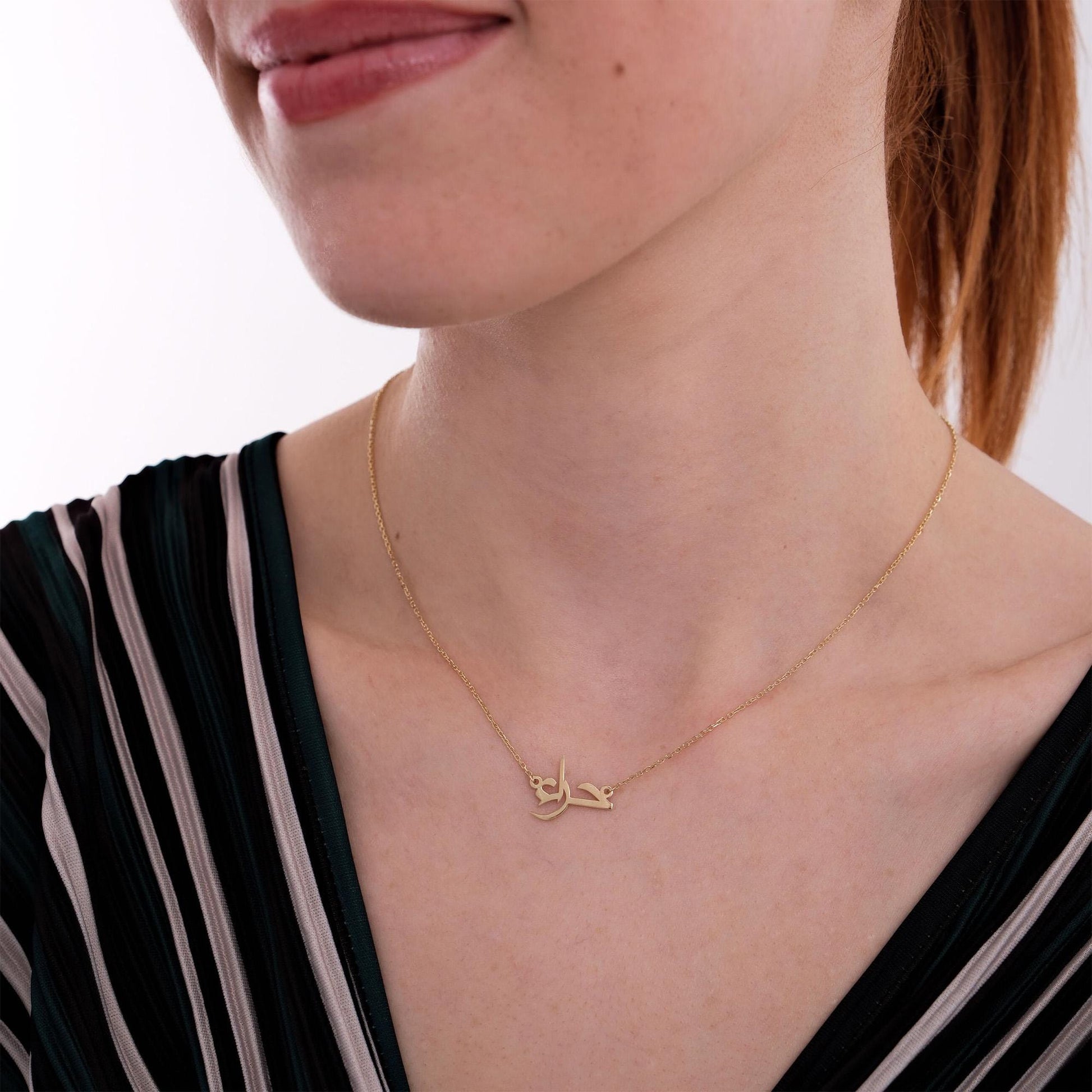 A woman smiling softly while wearing a 14K solid gold Arabic name necklace, the elegant pendant resting on her collarbone and complementing her striped V-neck top.