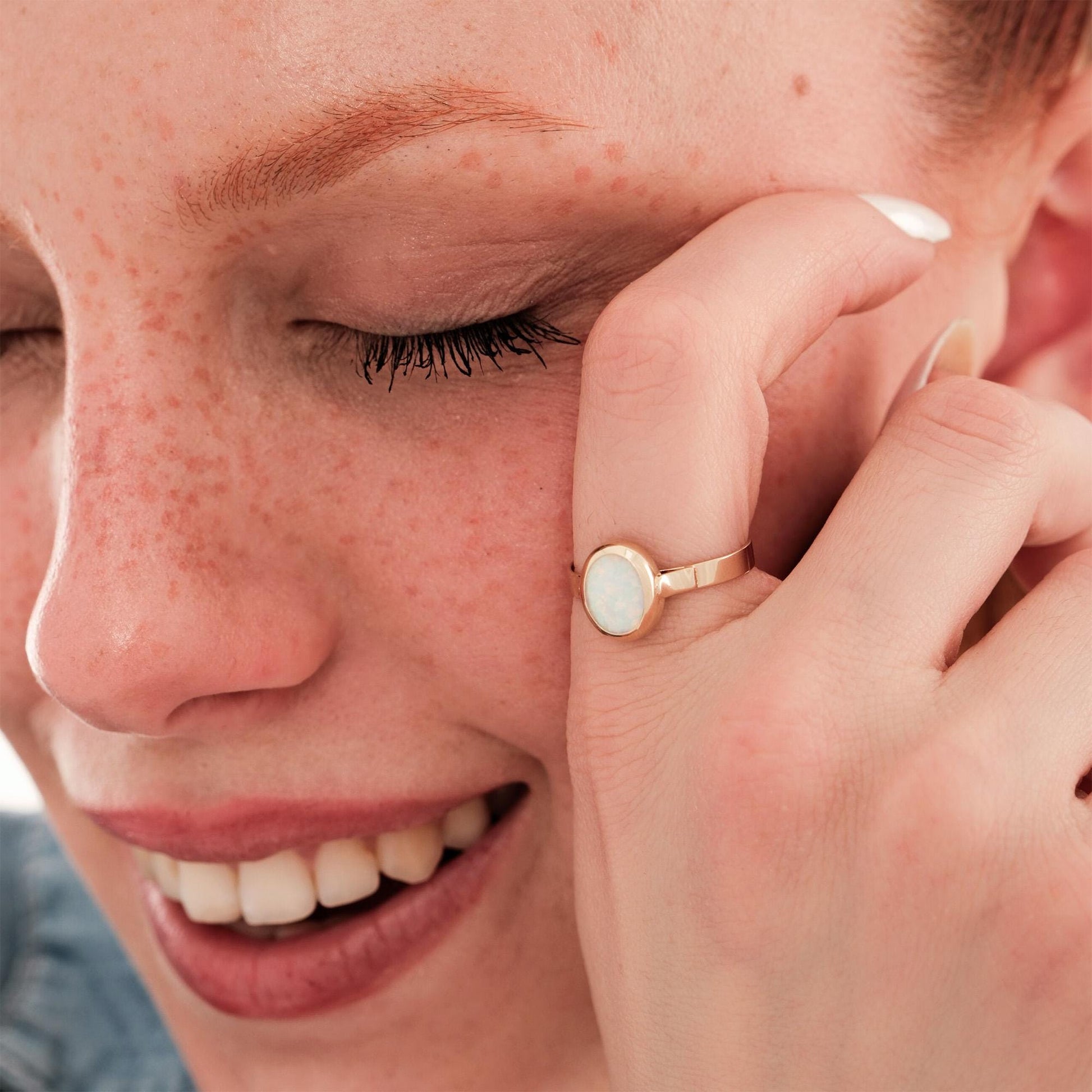A smiling woman with her eyes closed, wearing a 14K solid gold white opal ring that adds a soft glow and elegance to her natural look.