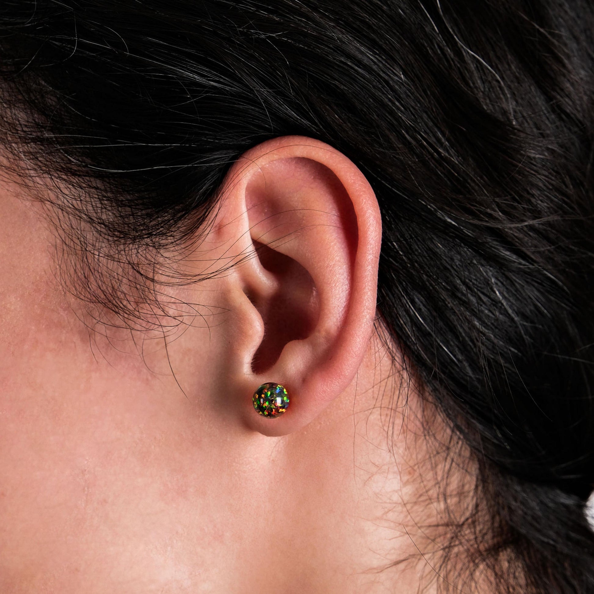 A woman with dark, wavy hair wearing fire opal stud earrings that reflect shades of green, red, and gold, set in 14K solid gold