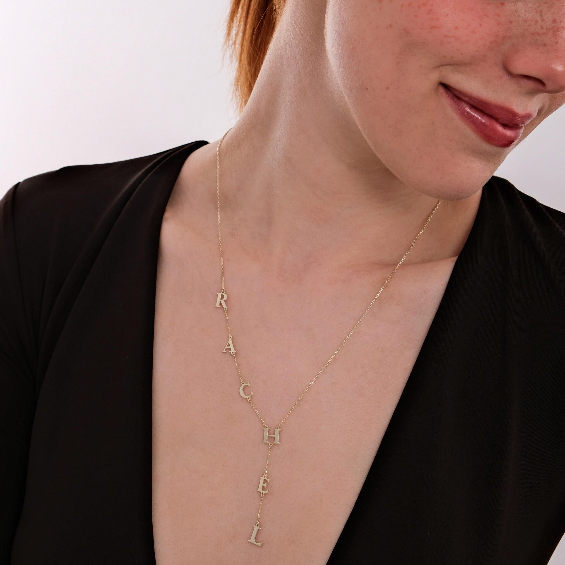 Smiling woman showing off a 14K solid gold personalized lariat necklace, styled with natural makeup and a low-cut black dress.