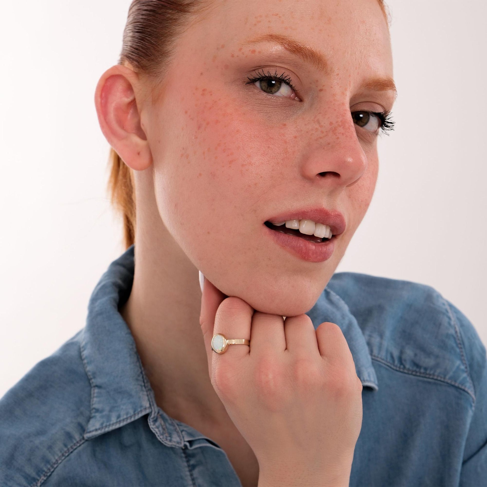Woman posing with a relaxed expression, showing off a 14K solid gold white opal ring on her index finger while wearing a denim shirt.