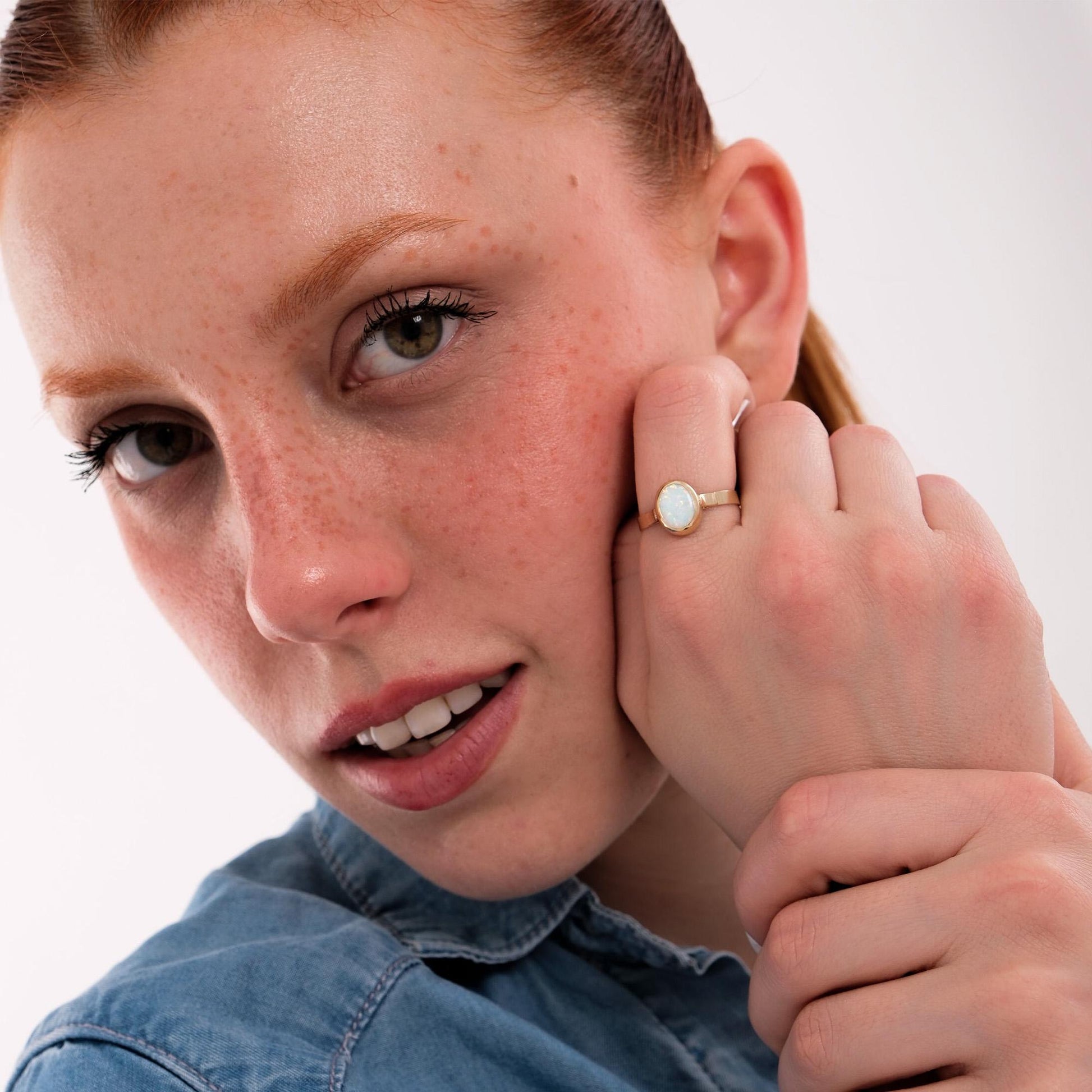 Woman resting her hand on her cheek, wearing a 14K solid gold white opal ring that beautifully complements her freckled skin and denim shirt.