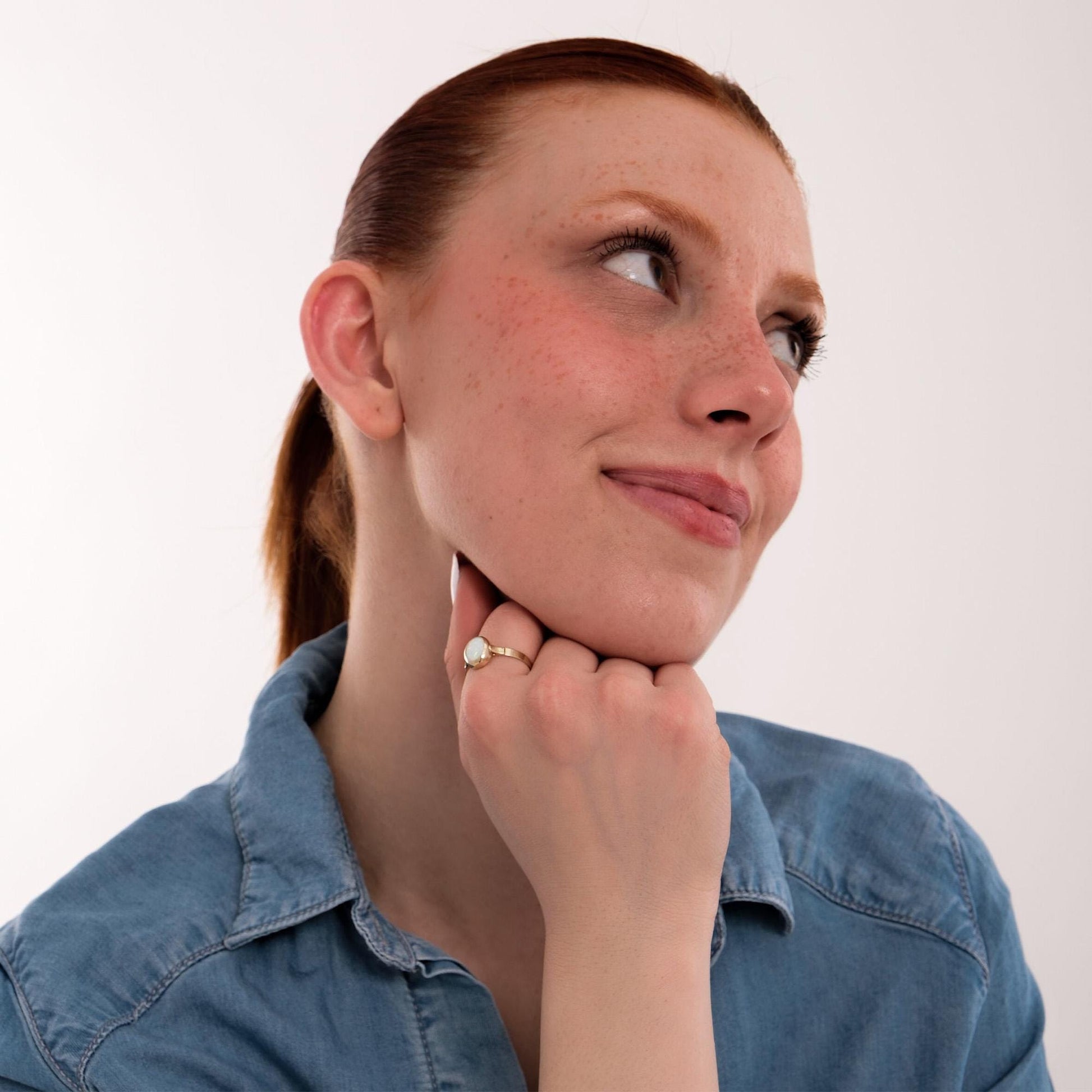 Woman looking up thoughtfully, wearing a 14K solid gold white opal ring that catches the light and adds a delicate sparkle to her casual outfit.