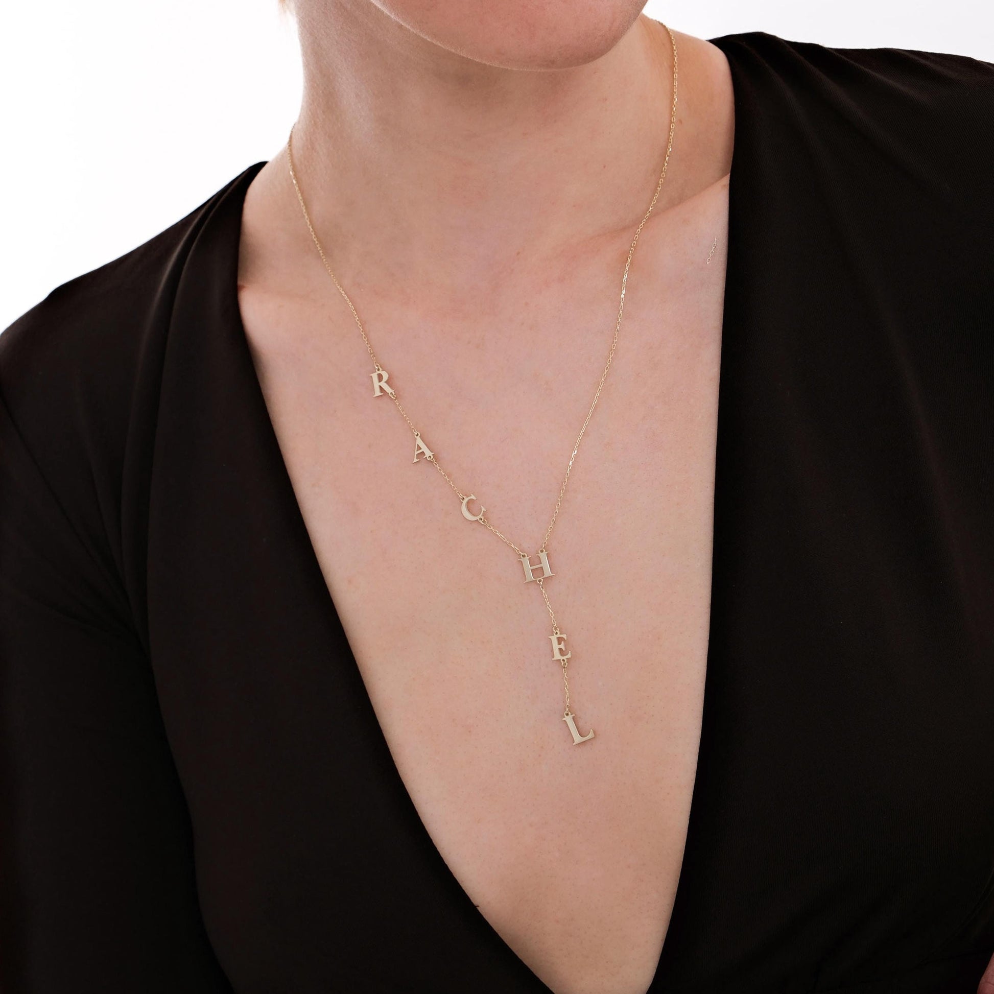 Woman wearing a custom 14K solid gold lariat necklace spelling “RACHEL”, shown from the front in an elegant black outfit.