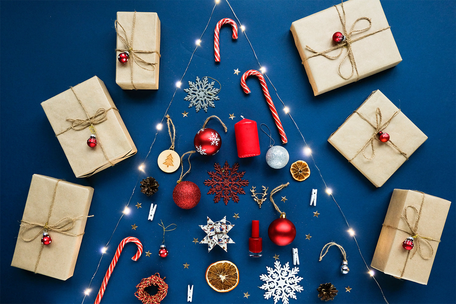 Christmas-themed arrangement on a blue background featuring wrapped gifts, candy canes, ornaments, and decorative items. The items are laid out in the shape of a Christmas tree, with lights outlining the tree shape. The gifts are wrapped in brown paper an