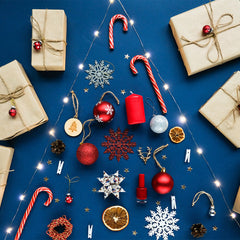 Christmas-themed arrangement on a blue background featuring wrapped gifts, candy canes, ornaments, and decorative items. The items are laid out in the shape of a Christmas tree, with lights outlining the tree shape. The gifts are wrapped in brown paper an