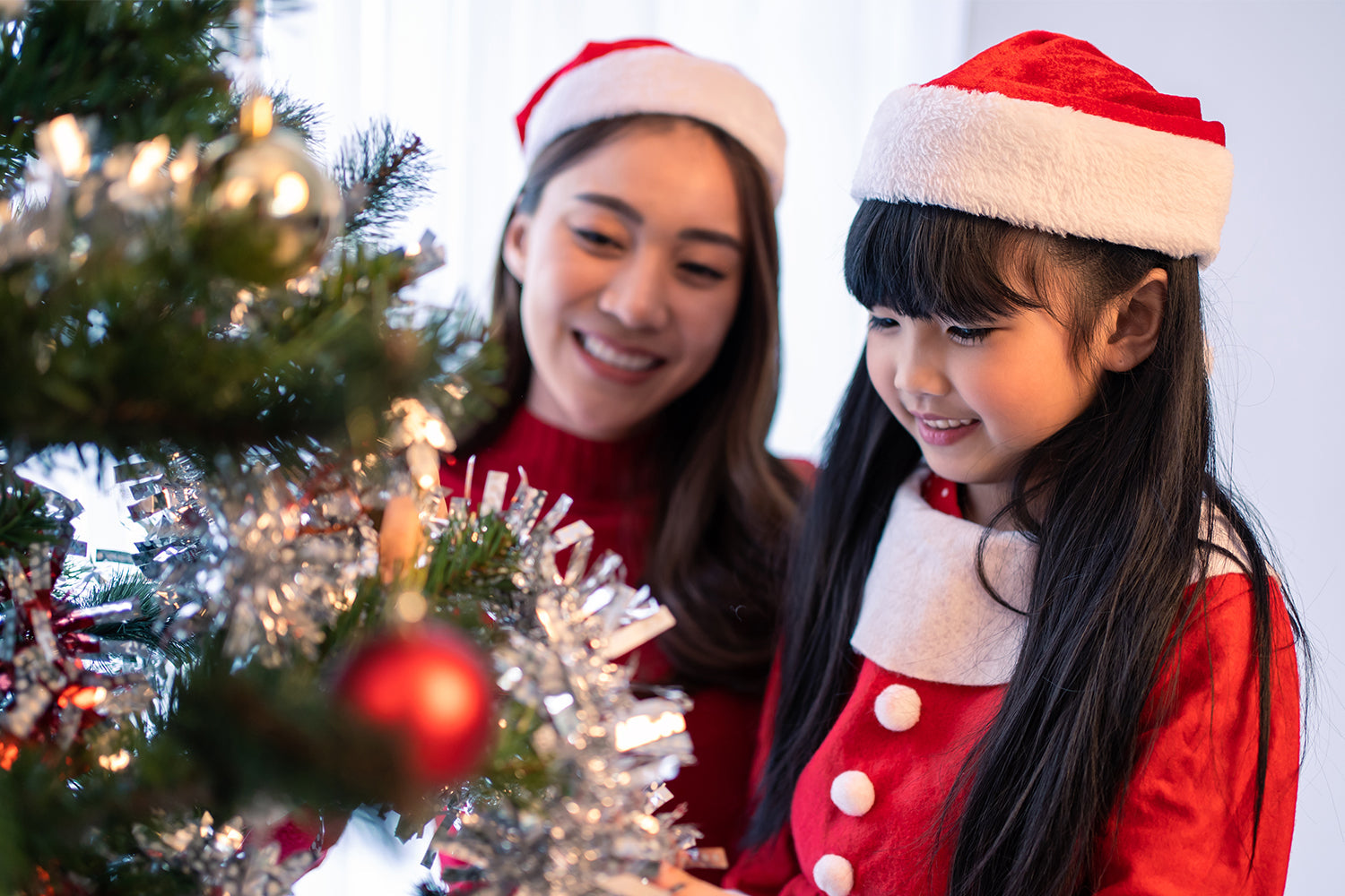 A mother and daughter, both wearing red Santa hats and matching red dresses with white trim, decorate a Christmas tree together. The mother smiles as she watches her daughter place an ornament on the tree. The tree is adorned with tinsel, lights, and red 