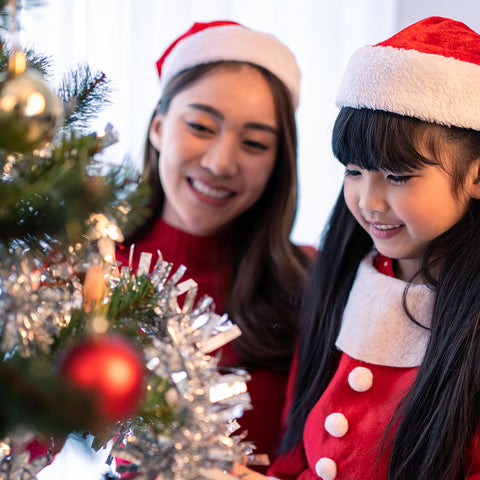 A mother and daughter, both wearing red Santa hats and matching red dresses with white trim, decorate a Christmas tree together. The mother smiles as she watches her daughter place an ornament on the tree. The tree is adorned with tinsel, lights, and red 