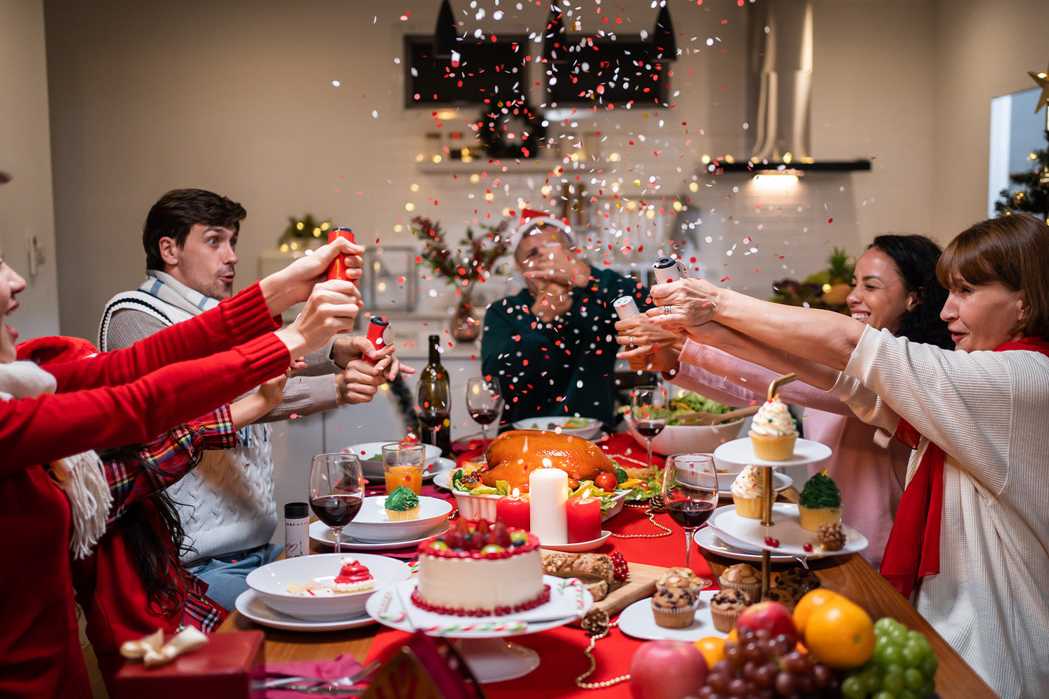 A multi-ethnic family gathered around a dinner table celebrating Christmas. They are pulling Christmas crackers, releasing red and white confetti. The table is adorned with a festive spread including a roasted turkey, a cake, cupcakes, and various fruits.