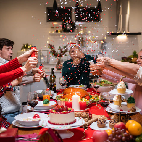 A multi-ethnic family gathered around a dinner table celebrating Christmas. They are pulling Christmas crackers, releasing red and white confetti. The table is adorned with a festive spread including a roasted turkey, a cake, cupcakes, and various fruits.