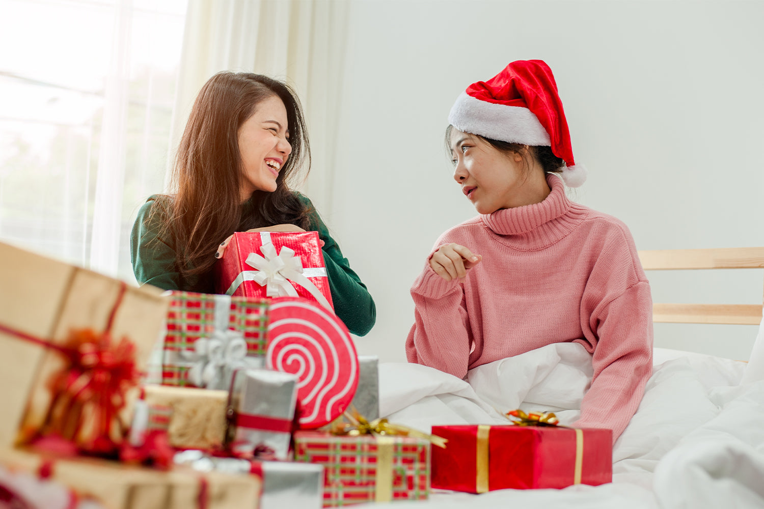 Two women sitting on a bed surrounded by Christmas gifts. One woman, wearing a green sweater, is laughing and holding a wrapped present with a white bow. The other woman, wearing a pink sweater and a Santa hat, looks at her with a smile. The bed is filled