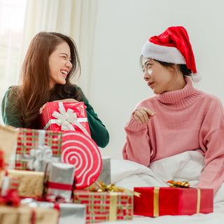 Two women sitting on a bed surrounded by Christmas gifts. One woman, wearing a green sweater, is laughing and holding a wrapped present with a white bow. The other woman, wearing a pink sweater and a Santa hat, looks at her with a smile. The bed is filled