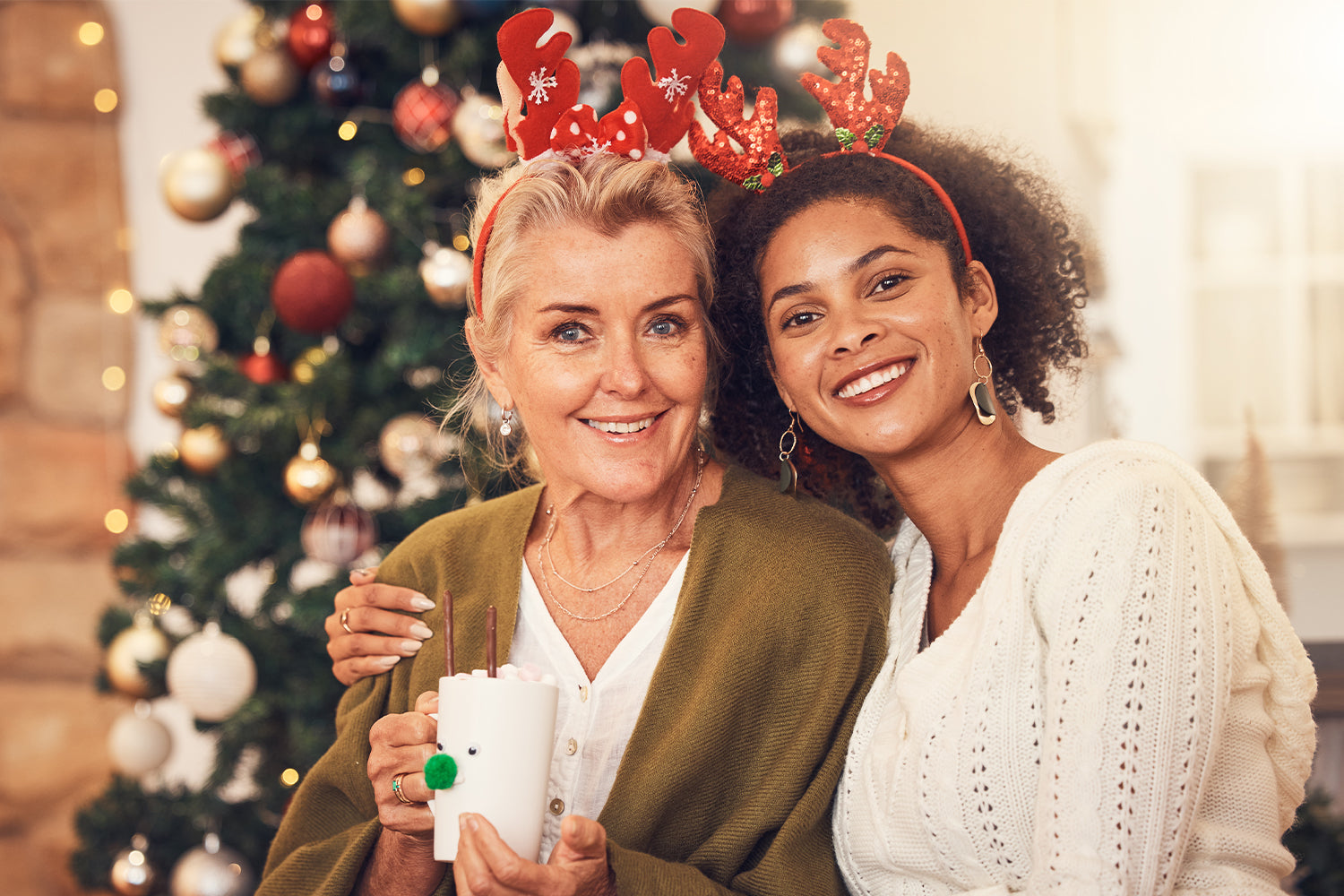 Two women, likely a mother and daughter, smiling and sitting together in front of a Christmas tree. Both wear reindeer antler headbands. The older woman holds a festive mug decorated with a reindeer face, while the younger woman has her arm around her. Th