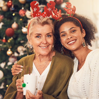 Two women, likely a mother and daughter, smiling and sitting together in front of a Christmas tree. Both wear reindeer antler headbands. The older woman holds a festive mug decorated with a reindeer face, while the younger woman has her arm around her. Th