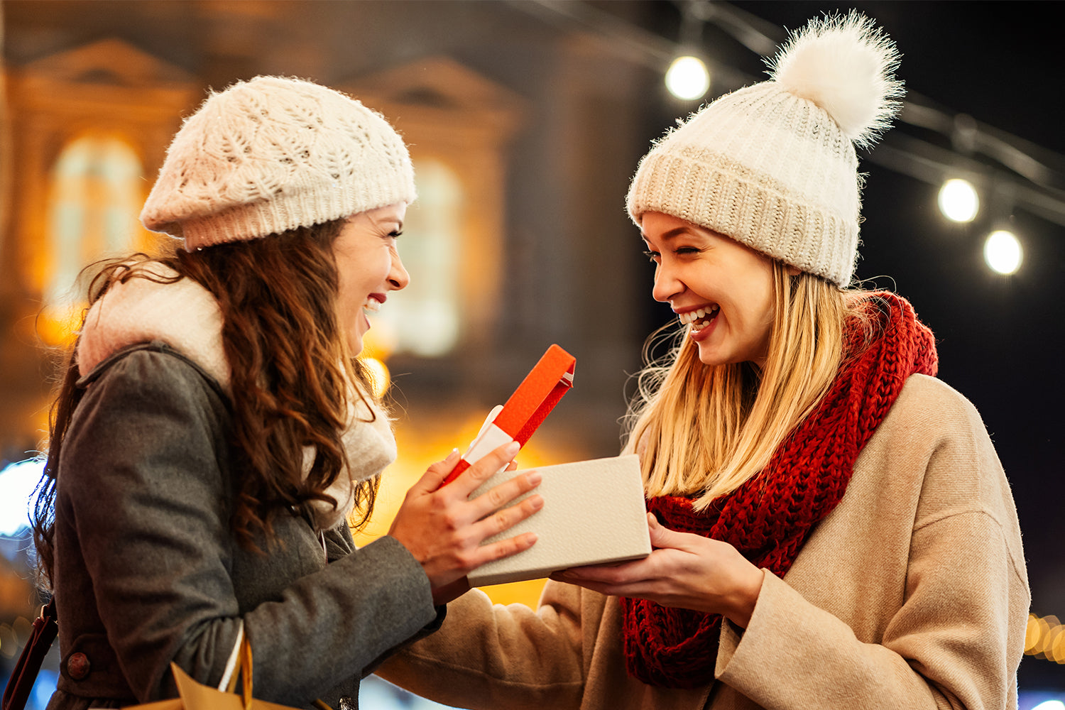 Two women wearing winter coats and knit hats exchanging Christmas gifts outdoors at night. One woman, holding a shopping bag, is opening a white gift box with a red lid while the other woman smiles and watches. Festive lights and blurred buildings in the 