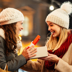 Two women wearing winter coats and knit hats exchanging Christmas gifts outdoors at night. One woman, holding a shopping bag, is opening a white gift box with a red lid while the other woman smiles and watches. Festive lights and blurred buildings in the 