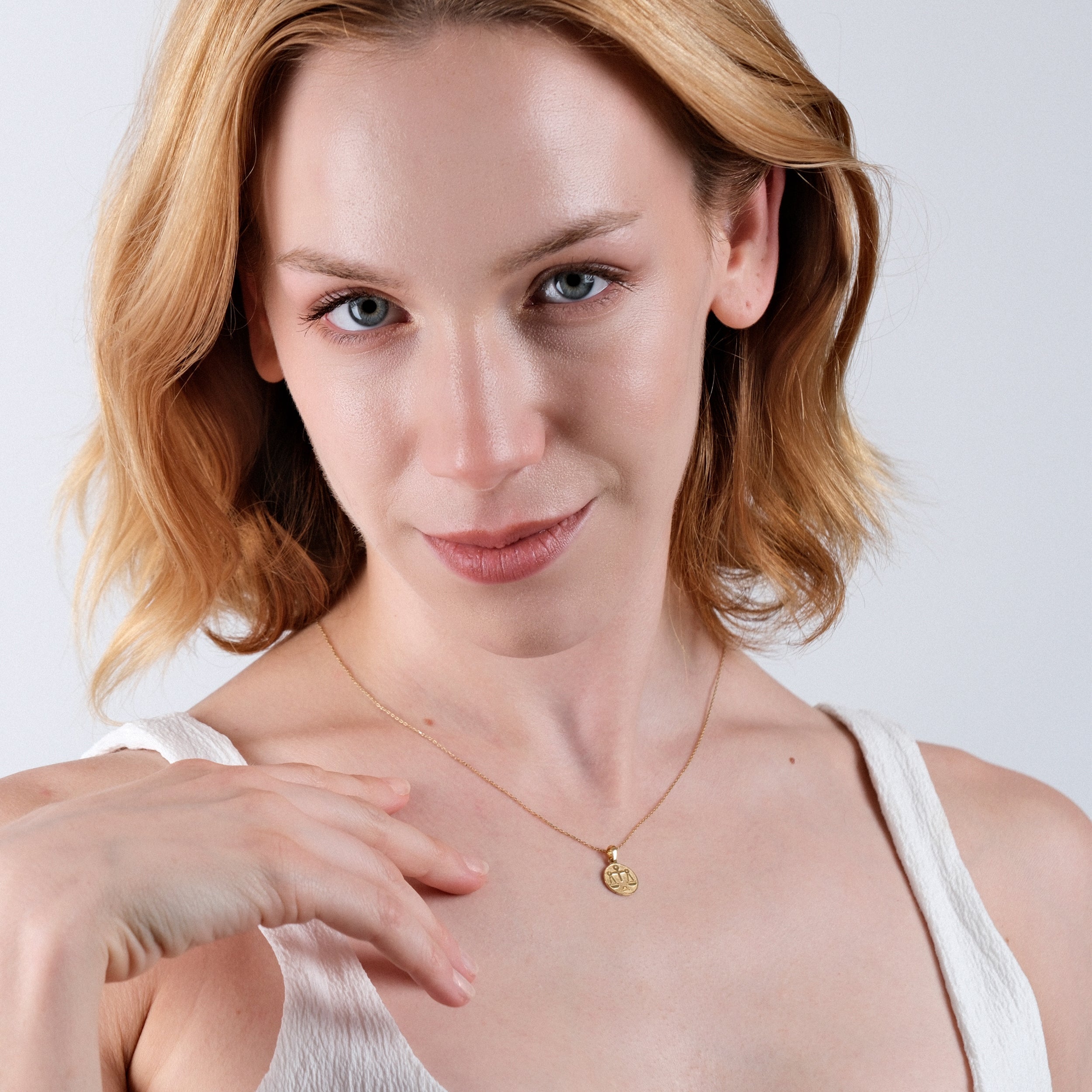Woman wearing a gold necklace with a pendant, short blonde hair, smiling, in a white top.