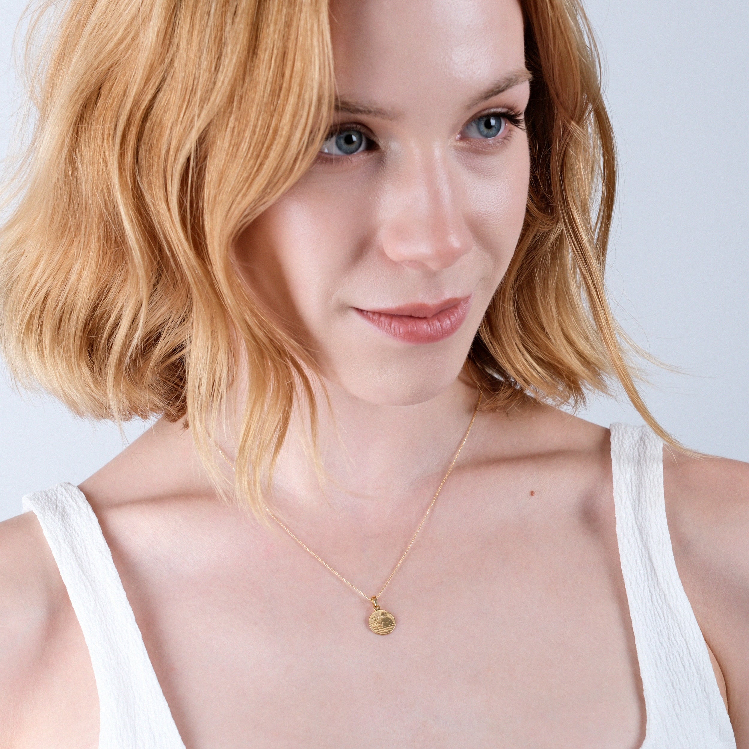 Woman with blonde hair wearing a delicate gold necklace and white top, smiling subtly against a neutral background.