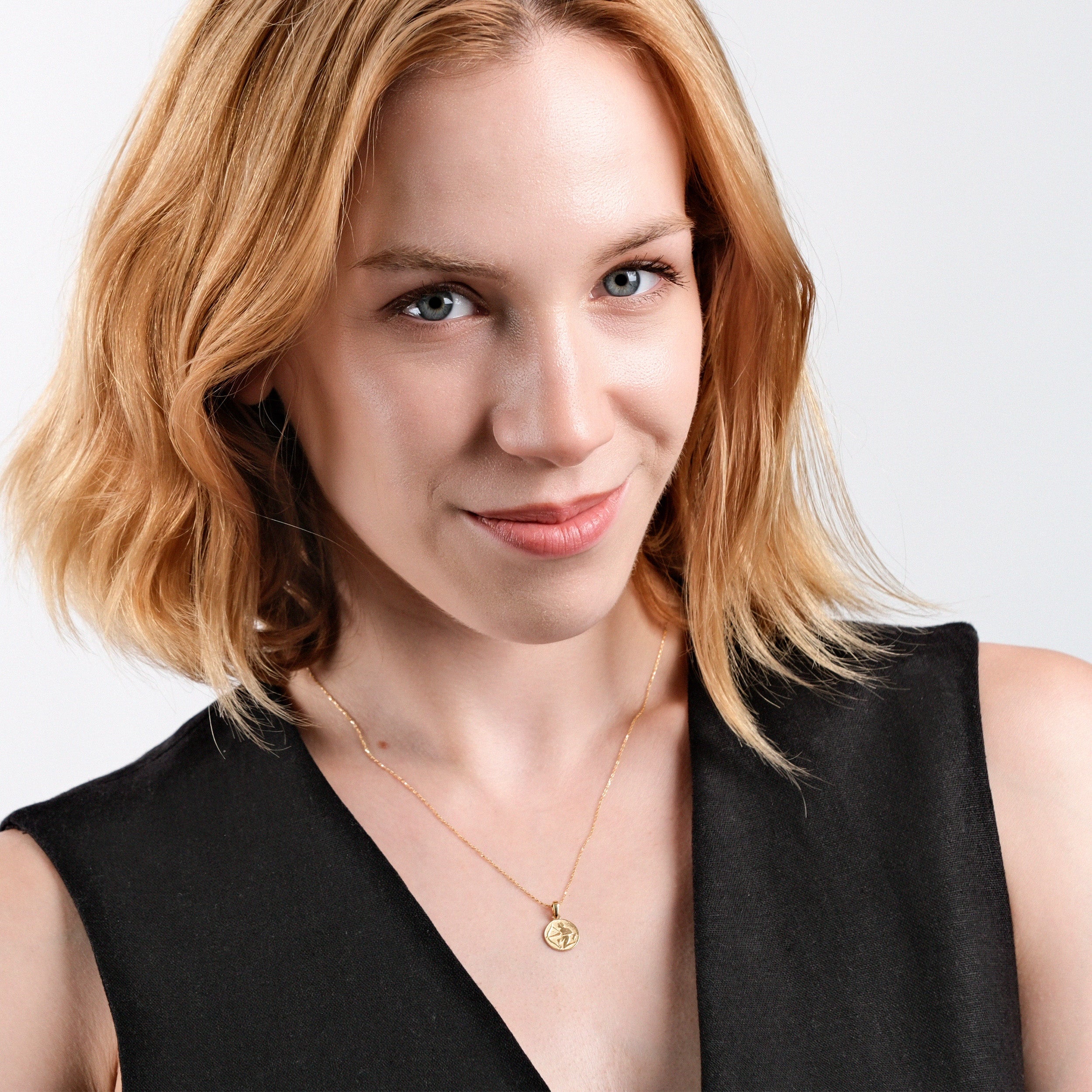 Smiling woman with short blonde hair wearing a gold necklace and black top against a white background.