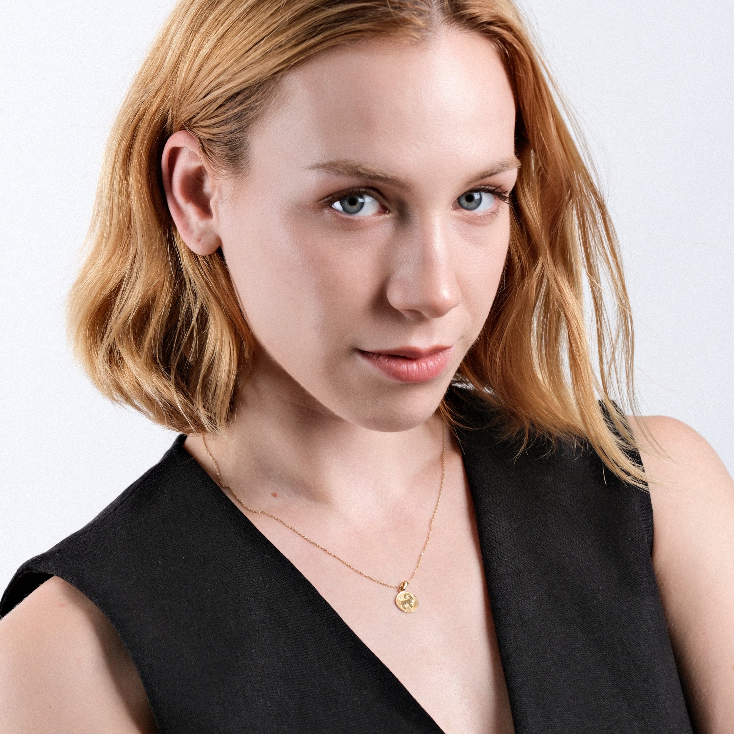 Blonde woman wearing black top and gold necklace, posing confidently with subtle smile on white background.