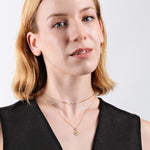 Woman wearing layered gold necklaces, short blonde hair, and black dress against white background, showcasing elegant jewelry.
