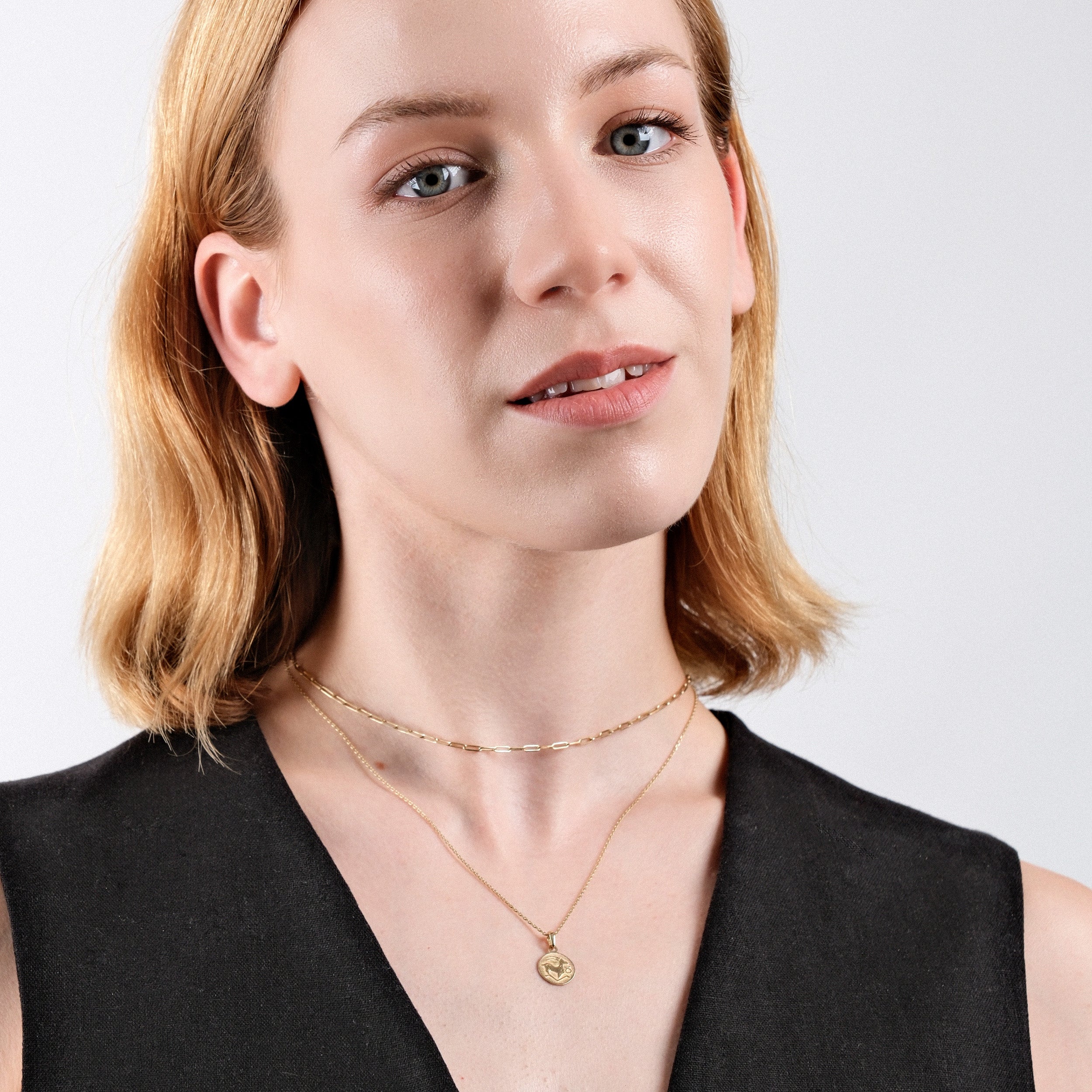 Woman wearing layered gold necklaces, short blonde hair, and black dress against white background, showcasing elegant jewelry.