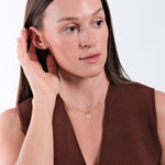Woman wearing gold necklace, brown top, touching ear, against white background, showcasing minimalist elegance.