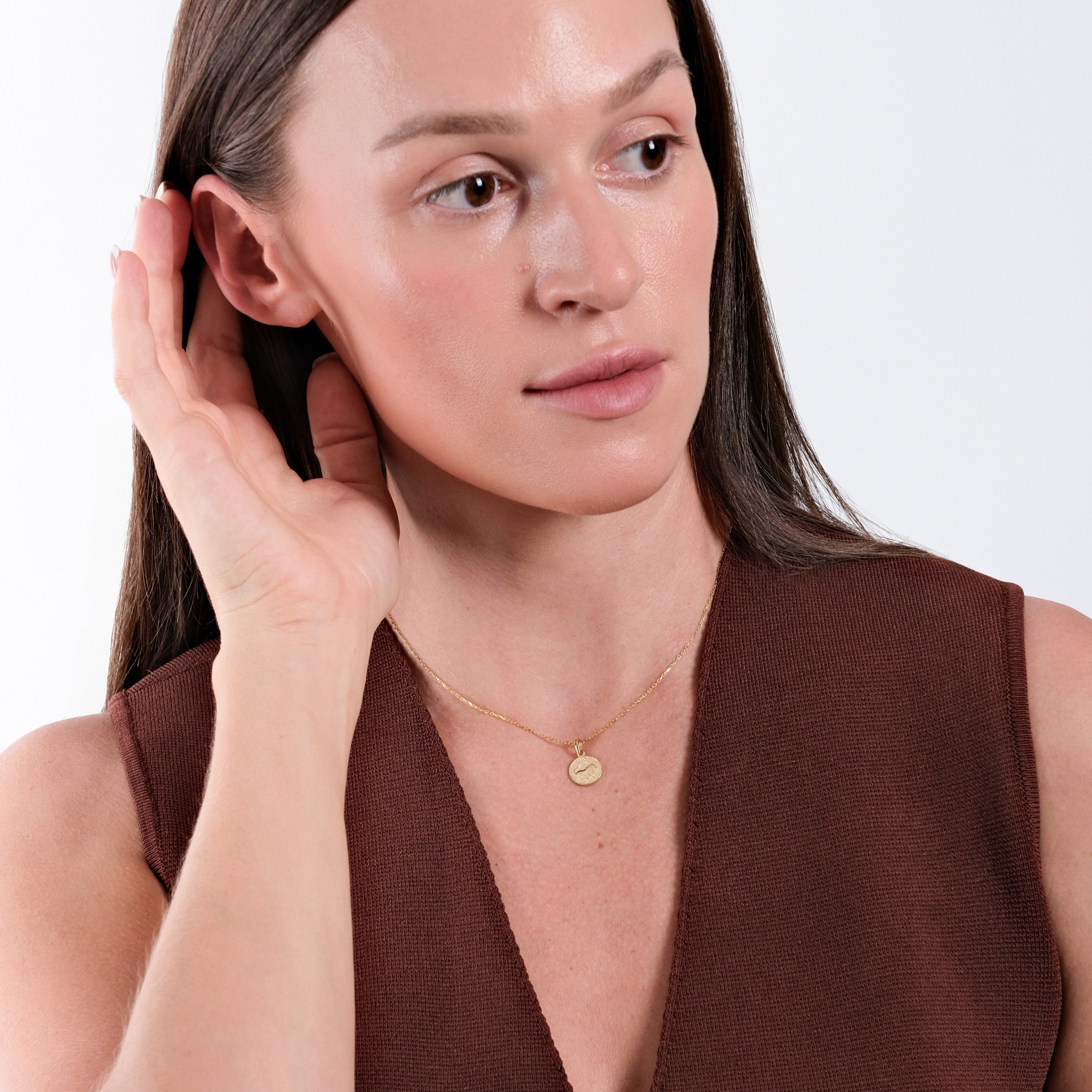 Woman wearing gold necklace, brown top, touching ear, against white background, showcasing minimalist elegance.