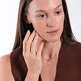 Woman with natural makeup wearing gold necklace and brown top, touching face gently with manicured hand.